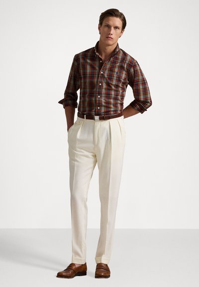 Chemise à carreaux boutonnée en rouge foncé, vert et jaune, associée à un pantalon beige à plis et des chaussures en cuir marron. Une ceinture marron complète le look.