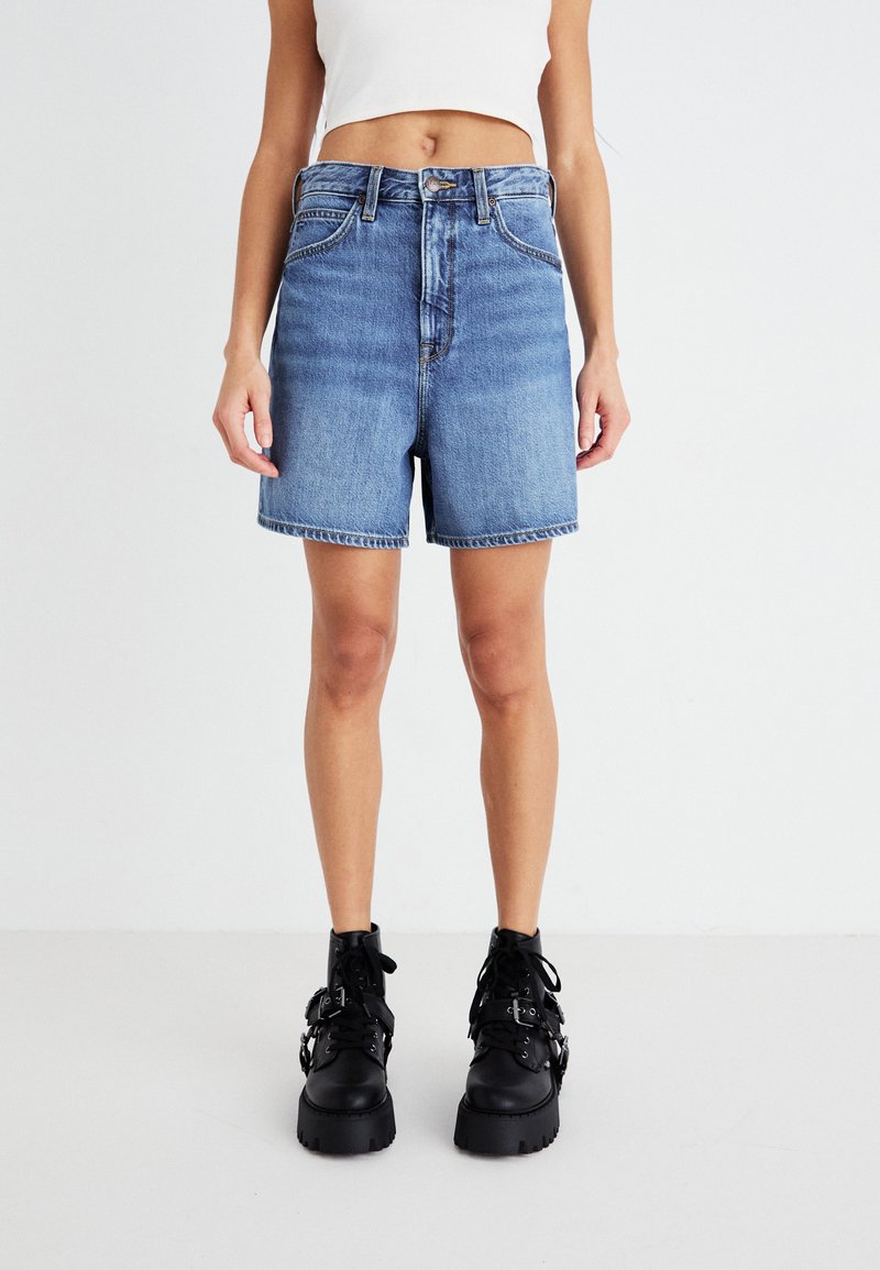 Shorts en denim bleu à taille haute, avec un ourlet effiloché et cinq poches, associés à des bottes noires à lacets épaisses avec des accents métalliques.