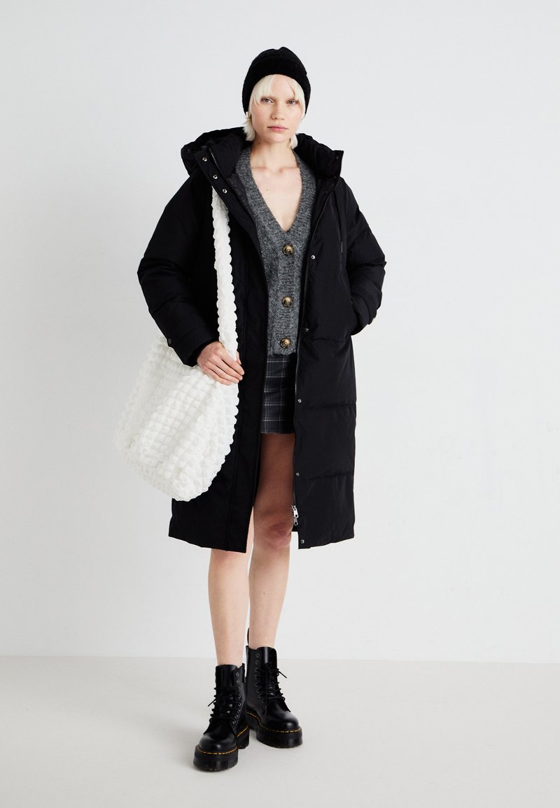 Schwarze Daunenjacke mit Kapuze, grauer Knopf-Down-Pullover, karierte Shorts und schwarze Schnürstiefel. Weiße strukturierte Tasche über der Schulter.