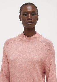 Maglione rosa lavorato a maglia con un collo alto a coste e maniche raglan. Texture morbida con una finitura leggermente melangiata.