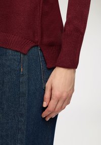 Bordowy sweter z dzianiny z prążkowanymi mankietami i bocznym rozcięciem, w komplecie z ciemnoniebieską spódnicą dżinsową. Sweter ma teksturowaną powierzchnię.