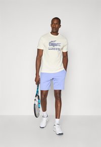T-shirt de manga curta em tom claro com logo gráfico azul, combinado com calções roxos claros. O modelo segura uma raquete de ténis e usa sapatilhas brancas.