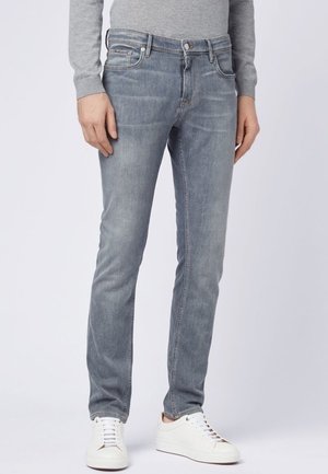 Persona vestida con vaqueros ajustados de color gris claro, camiseta de manga larga gris y zapatillas blancas, de pie frente a un fondo claro y liso.