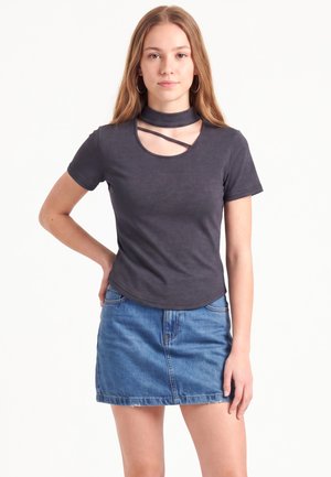 Jeune femme aux longs cheveux châtain clair portant un haut gris foncé à manches courtes avec un col découpé et une mini-jupe en jean bleue, debout devant un fond blanc.