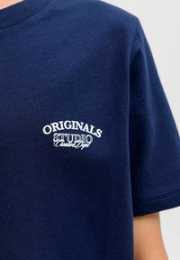 Navyblauwe katoenen T-shirt met witte geborduurde tekst "ORIGINALS STUDIO" op de linkerborst. Korte mouwen en ronde hals.