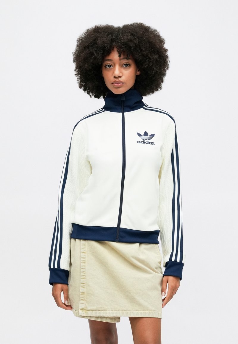 White zip-up jacket with navy accents and three stripes on sleeves. Features a high collar and embroidered logo on the chest. Soft texture.