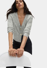 Cardigan in grigio chiaro, lavorato a maglia, con scollo a V, caratterizzato da tre bottoni dorati e polsini a coste. Indossato sopra pantaloni scuri.