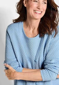 Heller blauer strukturierter Cardigan mit gepunktetem Muster, gewelltem Ausschnitt und Knopfschluss. Lange Ärmel und weiches Strickmaterial.