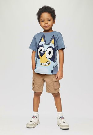 Blaues Baumwoll-T-Shirt mit Druck eines Cartoon-Hundegesichts, kombiniert mit beige Cargo-Shorts. Das Kind trägt weiße Sneaker mit grauen Akzenten.