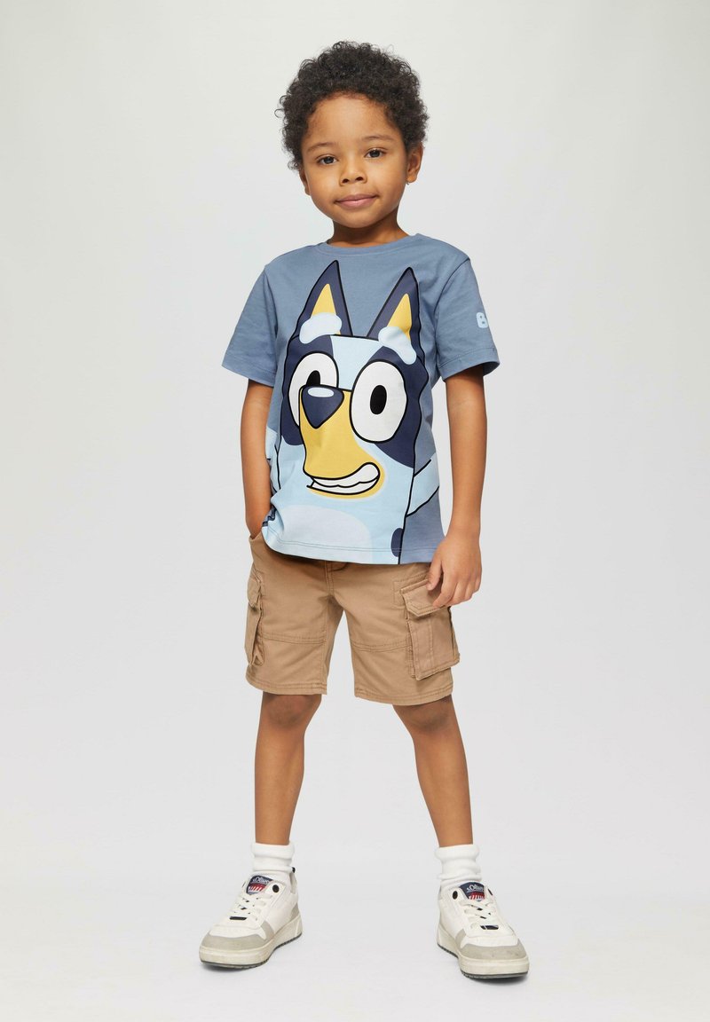 Blauwe katoenen t-shirt met een print van een cartoonhondengezicht, gecombineerd met zandkleurige cargo shorts. Het kind draagt witte sneakers met grijze accenten.