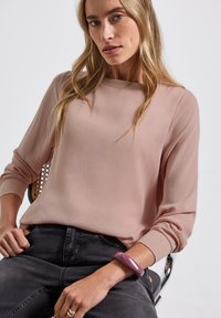 Blush blouse met lange mouwen, een gladde textuur en brede manchetten, gecombineerd met donkere spijkerbroeken. Het model draagt een paarse armband om haar pols.