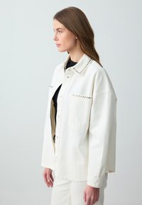 Veste en denim blanche avec des clous métalliques le long du col et de la poche. Dotée d'une coupe décontractée, d'une fermeture à boutons à l'avant et de manches longues.