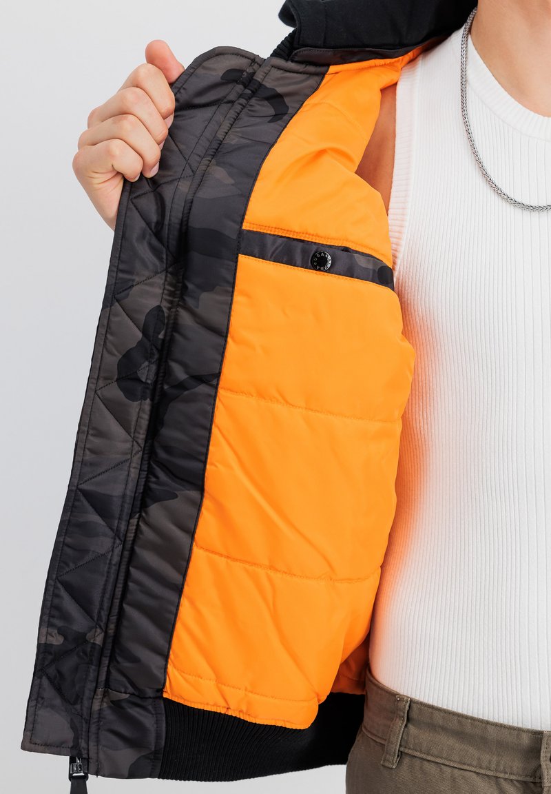 Giacca trapuntata nera con una fodera isolante arancione e una tasca a toppa; rifinita con polsini a costine neri.