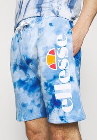 Shorts tie-dye bleus avec une taille élastique, arborant le logo blanc "ellesse" et un design d'accent coloré. Matière douce en mélangé de coton.