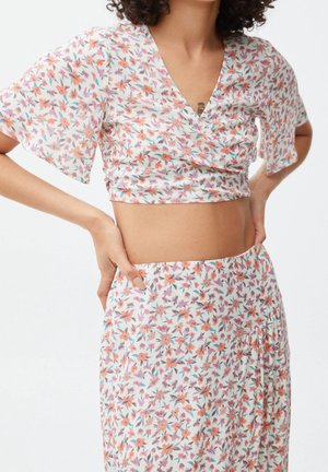 Vrouw in een witte bloeiende crop top met vlindermouwen en een bijpassende hoge taille rok, met haar handen op de heupen, tegen een witte achtergrond.