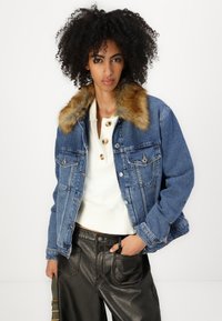 Denim jacket with a brown faux fur collar, two chest pockets, and orange stitching. Worn over a white, buttoned sweater and black leather pants.