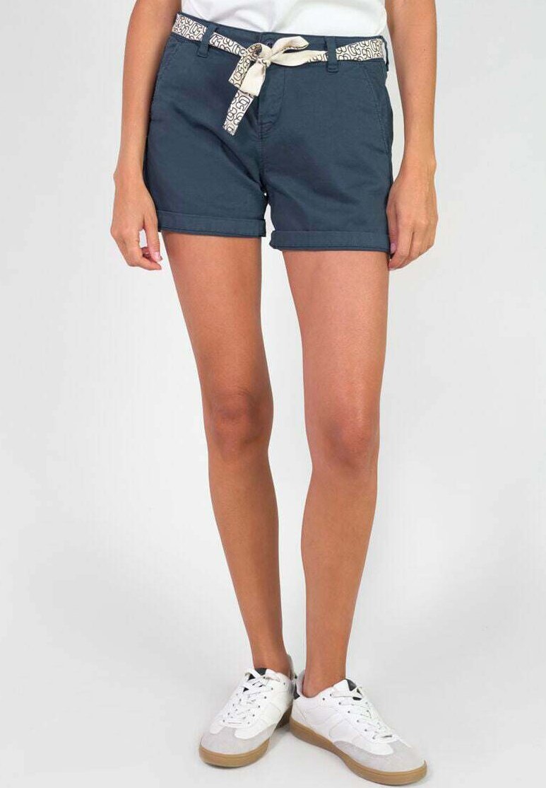 Navyblauwe katoenen shorts met een opgerolde zoom, voorzien van een gestreepte crèmekleurige riem. Draagt witte sneakers, wat de casual zomerse stijl benadrukt.