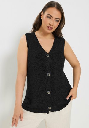 Vrouw met een zwarte gebreide mouwloze knoopvest en lichtgekleurde broek, poserend met een hand op haar heup tegen een effen achtergrond.