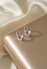 Silver hoop örhängen med en modern design, som har en slät yta och en distinkt rund form, vilande på en beige satin yta.