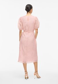 Kvinna med hår i bulle klädd i en rosa midiklänning med puffärmar och guldiga högklackade sandaler, stående med ryggen mot kameran.