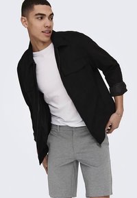 Juoda striukė su užtrauktuku ir krūtinės kišenėmis, dėvima virš balto marškinėlio ir pilkų šortų. Audinys atrodo lygus, su laisvu prigludimu.