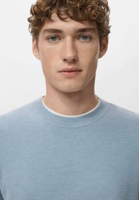 Jasnoniebieski dziergany sweter z okrągłym dekoltem. Teksturowana tkanina ma wzór plastra miodu. Modelka ma jasną cerę i krótkie kręcone włosy.