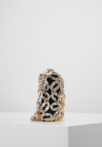 Czarna torebka owinięta dużymi, splecionymi złotymi i srebrnymi łańcuchami, wyeksponowana na białym piedestale na jednolitym tle.