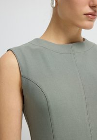 Zelené šaty bez rukávů s vysokým výstřihem, švy a hladkou texturou. Žádné viditelné zapínání nebo vzory.