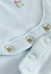 Helles blaues Gewebe mit kleinen Blumenstickereien, silbernen Druckknöpfen und Knopfdetails an Kragen und Bündchen. Texturiertes Strickmaterial.