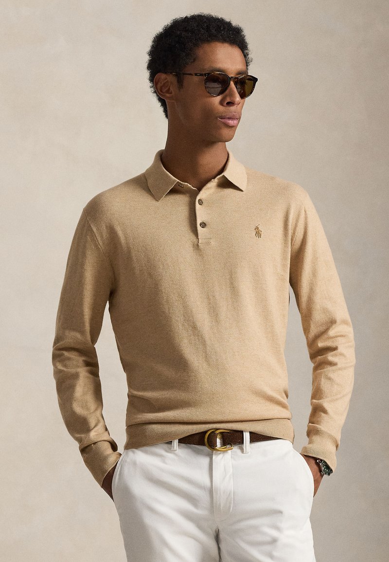 Polo a maniche lunghe beige con colletto, tre bottoni e logo ricamato. Abbinata a pantaloni bianchi e cintura marrone con fibbia dorata.