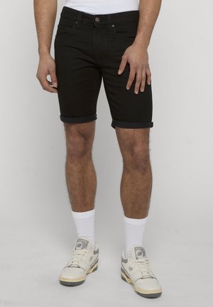 Mann iført svarte oppbrettede denimshorts, hvite crew-sokker, hvite og grå New Balance-sneakers, står mot en ensfarget bakgrunn.