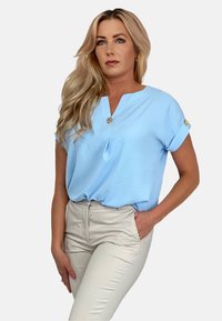 Jasnoniebieska bluzka z dekoltem w serek, krótkimi podwijanymi rękawami i ozdobnym detalem na guziku. Noszona z dopasowanymi spodniami w kolorze kremowym.