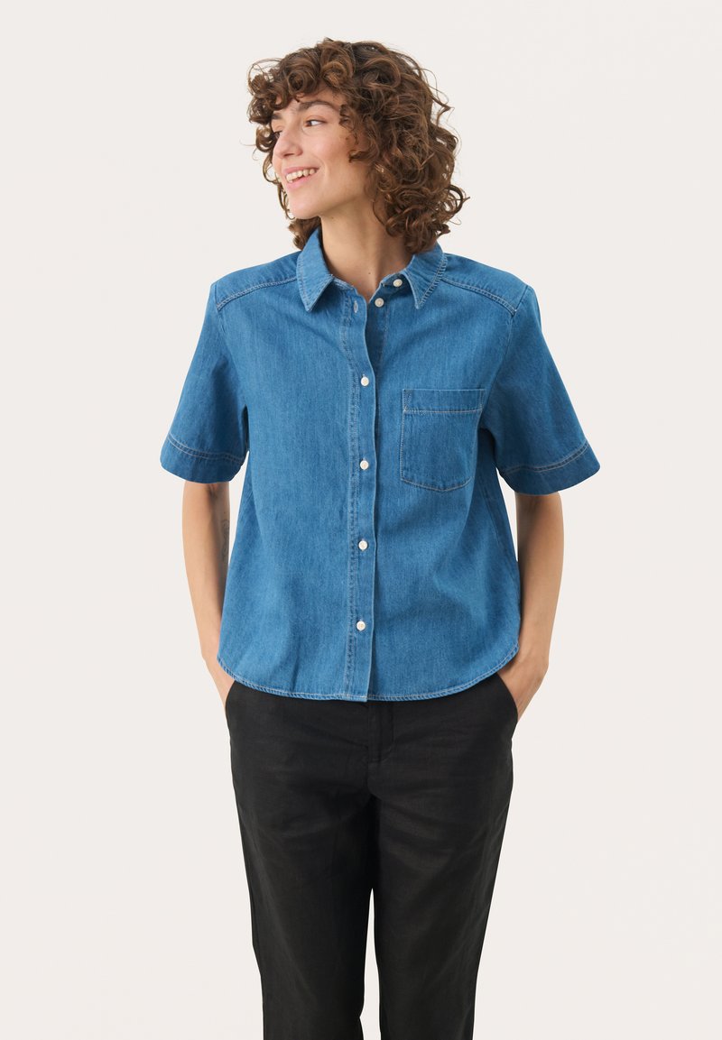 Camicia di jeans blu a maniche corte con chiusura a bottoni, taschino, e orlo curvo. Abbinata a pantaloni neri, di texture liscia.
