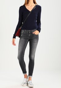 Suéter de pico azul marino, jeans negros ajustados con detalles desgastados, zapatos plateados tipo slip-on y un bolso pequeño rojo con correa.