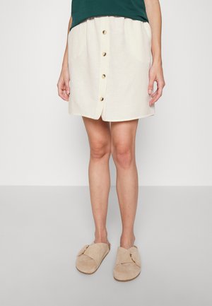 Saia mini em mistura de linho em creme, com fecho frontal por botões e dois bolsos laterais. Combinada com chinelos bege para um look casual.