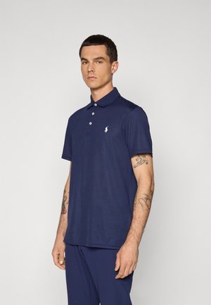 Mann i marineblå polo med en hvit brodert logo, mørke bukser og synlige tatoveringer på begge underarmer, står mot en ensfarget bakgrunn.