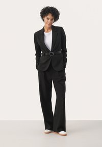 Costume noir sur mesure avec une veste ceinturée et un pantalon large. La veste présente un col cranté et des poches ; associée à des baskets blanches.