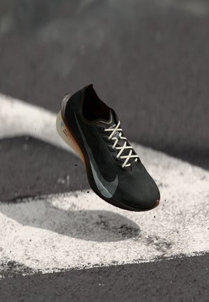 Czarny but sportowy z teksturowaną cholewką, białymi sznurówkami i szarym logo typu swoosh. Posiada amortyzowaną podeszwę i brązową wkładkę. W powietrzu, nad chodnikiem.