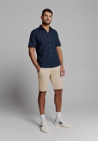 Navy korte mouwen button-up shirt, beige shorts, witte sneakers met blauwe accenten. Gladde stof, relaxte pasvorm, casual kleding voor warm weer.