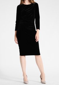 Svart sammetsklänning med rund halsringning, långa ärmar och rynkad detalj vid midjan, som faller precis under knäet.