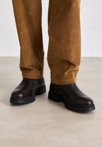 Stivali alla caviglia in pelle marrone con una texture liscia, punta rotonda e una robusta suola in gomma nera; abbinati a pantaloni in corduroy marroni.