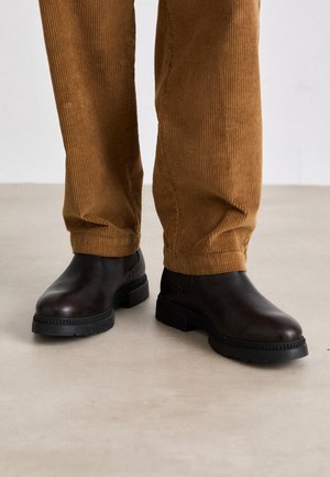 Bottines en cuir marron avec une texture lisse, bout rond et une épaisse semelle en caoutchouc noir ; accompagnées de pantalons en velours côtelé marron.
