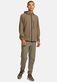 Man staat terwijl hij een bruin hooded jack, olijfgroene broek en bruin trailrunning schoenen met kleurrijke veters draagt, tegen een witte achtergrond.