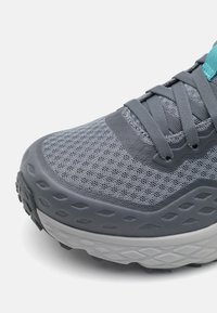 Sapatilha atlética cinza com parte superior em malha, sobreposição texturizada e detalhe em teal. Apresenta uma sola branca espessa com padrão para maior tração.