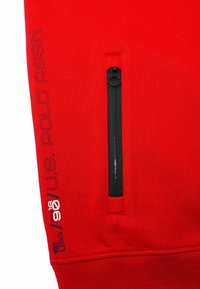Röd sweatshirttyg har en svart dragkedjeficka. På vänster ärm står "U.S. POLO ASSN." broderat i svart. Slät textur.