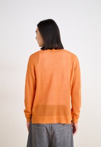 Oranges Strickpullover mit lockerer Passform, der ein strukturiertes Muster, gerippte Bündchen und einen abgerundeten Saum aufweist. Getragen mit grauen Shorts.