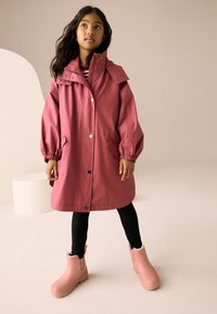 Rózsaszín vízálló kabát magas gallérral, patentgombokkal, oldalzsebekkel és rugalmas mandzsettákkal, rózsaszín bokacsizmával fekete leggingssel párosítva.
