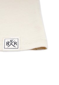 Beige puuvillakangas, jossa on huoliteltu reuna ja pieni kudottu etiketti, jossa on mustat kirjaimet ja ristiin menevät viivat. Sujuva pinta, ei näkyviä kuvioita.