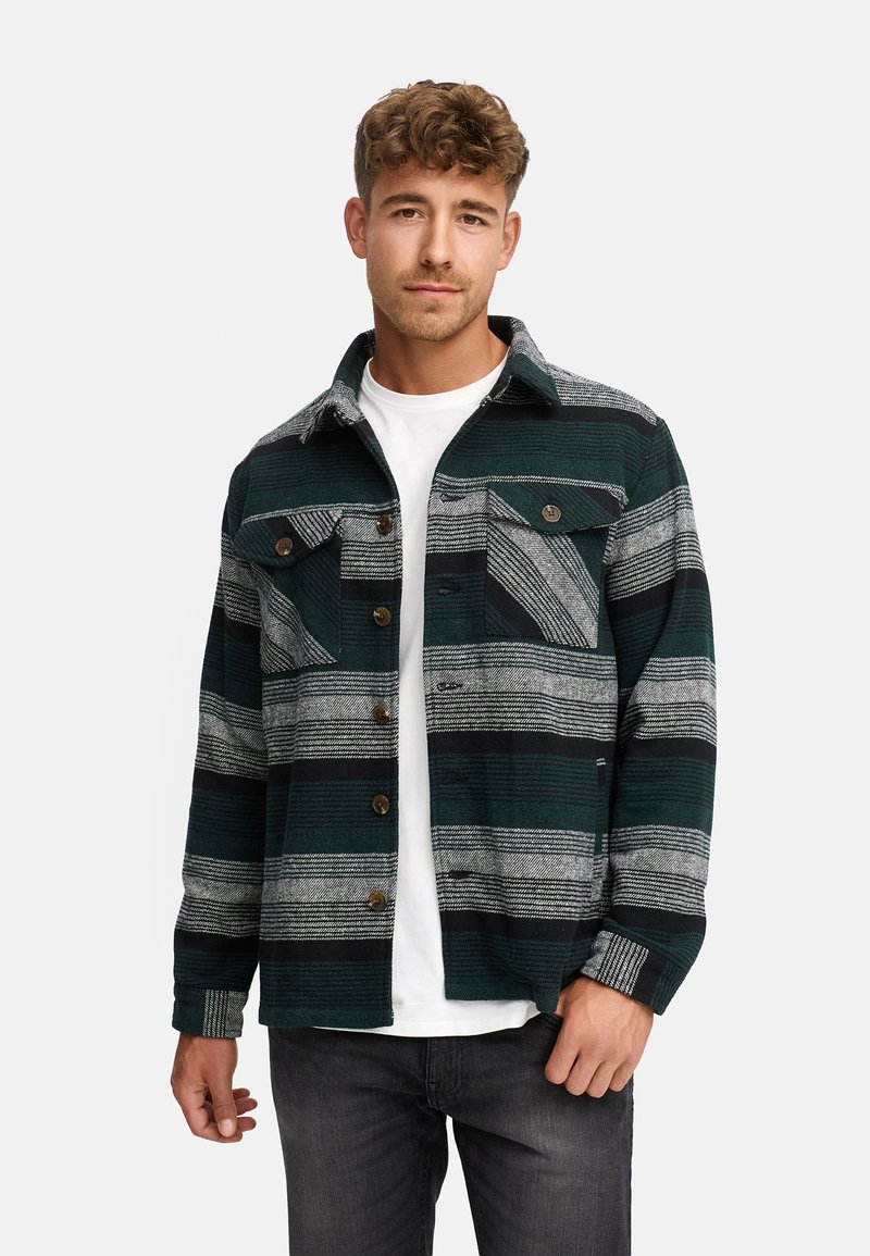 Grøn og grå stribet flanelskjorte med kontrasterende knapper og to brystlommer, båret over en hvid t-shirt, med en klassisk krave.