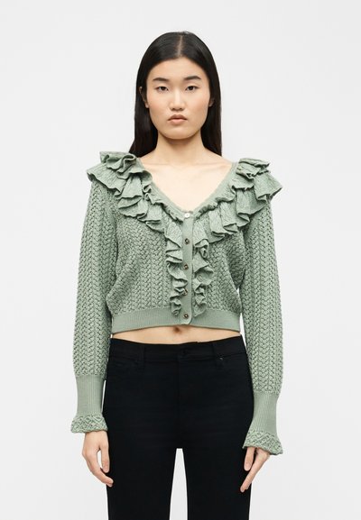 Woman wearing a cropped sage green knit cardigan with ruffles along the V-neck and front, paired with black high-waisted pants.
